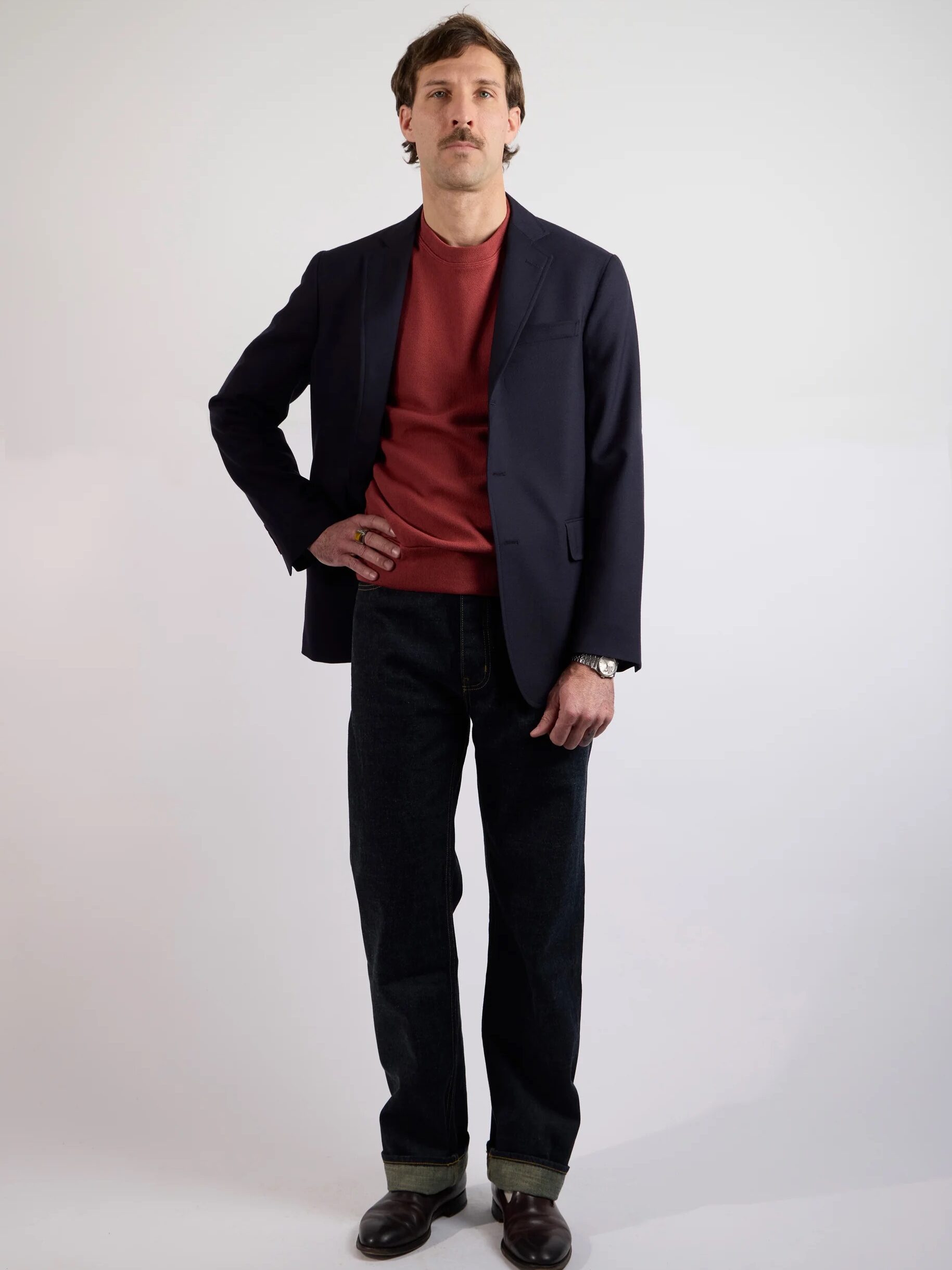 A man stands against a plain background wearing a navy blazer, red sweater, dark jeans, and brown shoes, with one hand in his pocket.