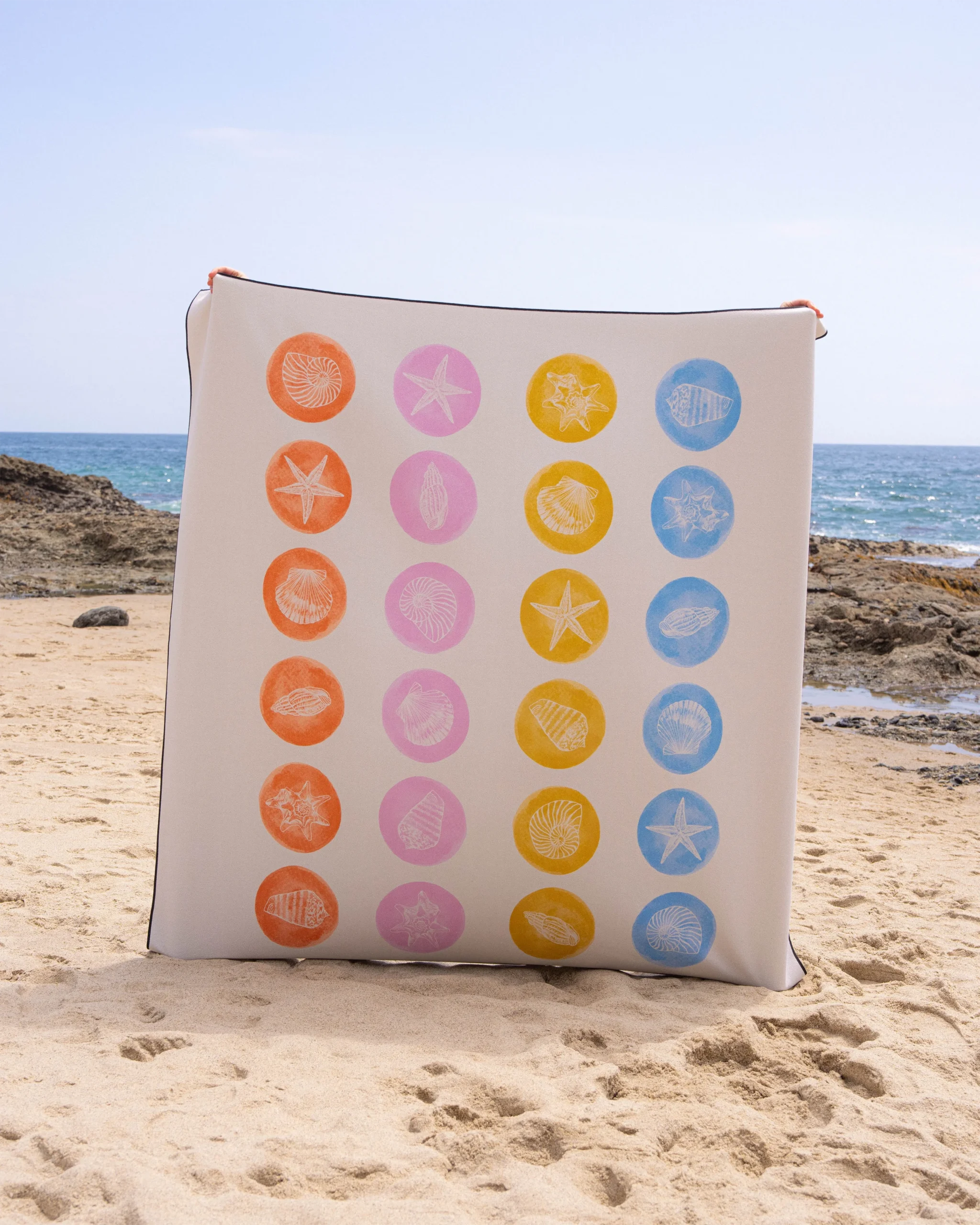 A square cushion with rows of colorful seashell and starfish prints stands upright on a sandy beach with rocks and the ocean in the background.