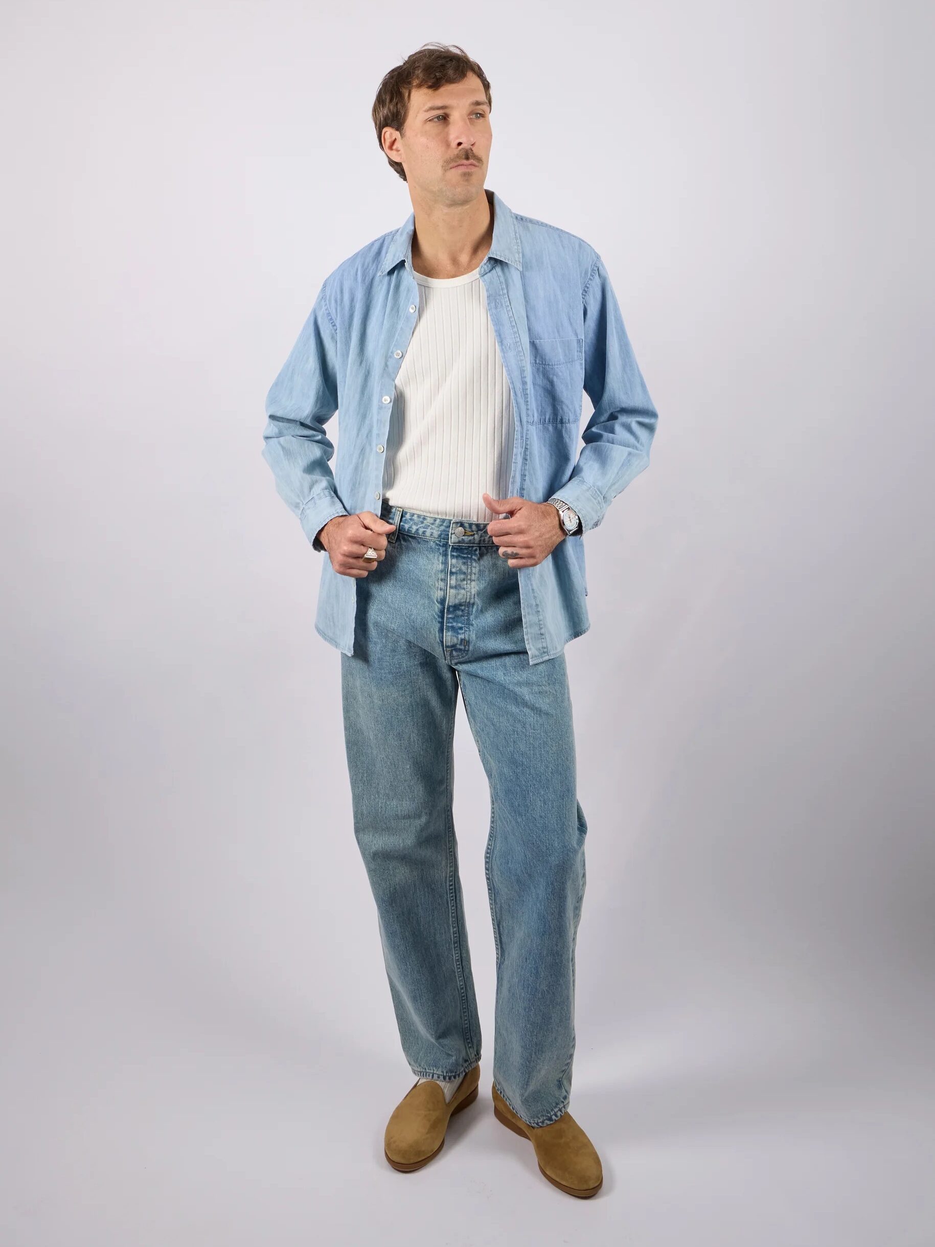 A man stands against a plain background wearing a light blue button-up shirt, white ribbed tank top, blue jeans, and brown shoes.