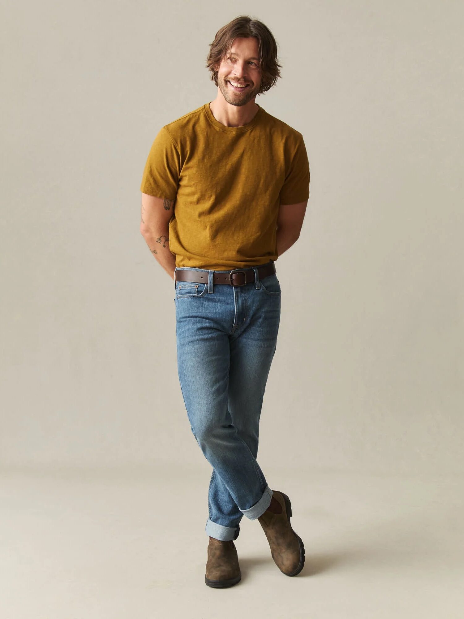 A man stands against a plain background, wearing a mustard t-shirt, blue cuffed jeans, brown belt, and brown boots, smiling with hands behind his back.