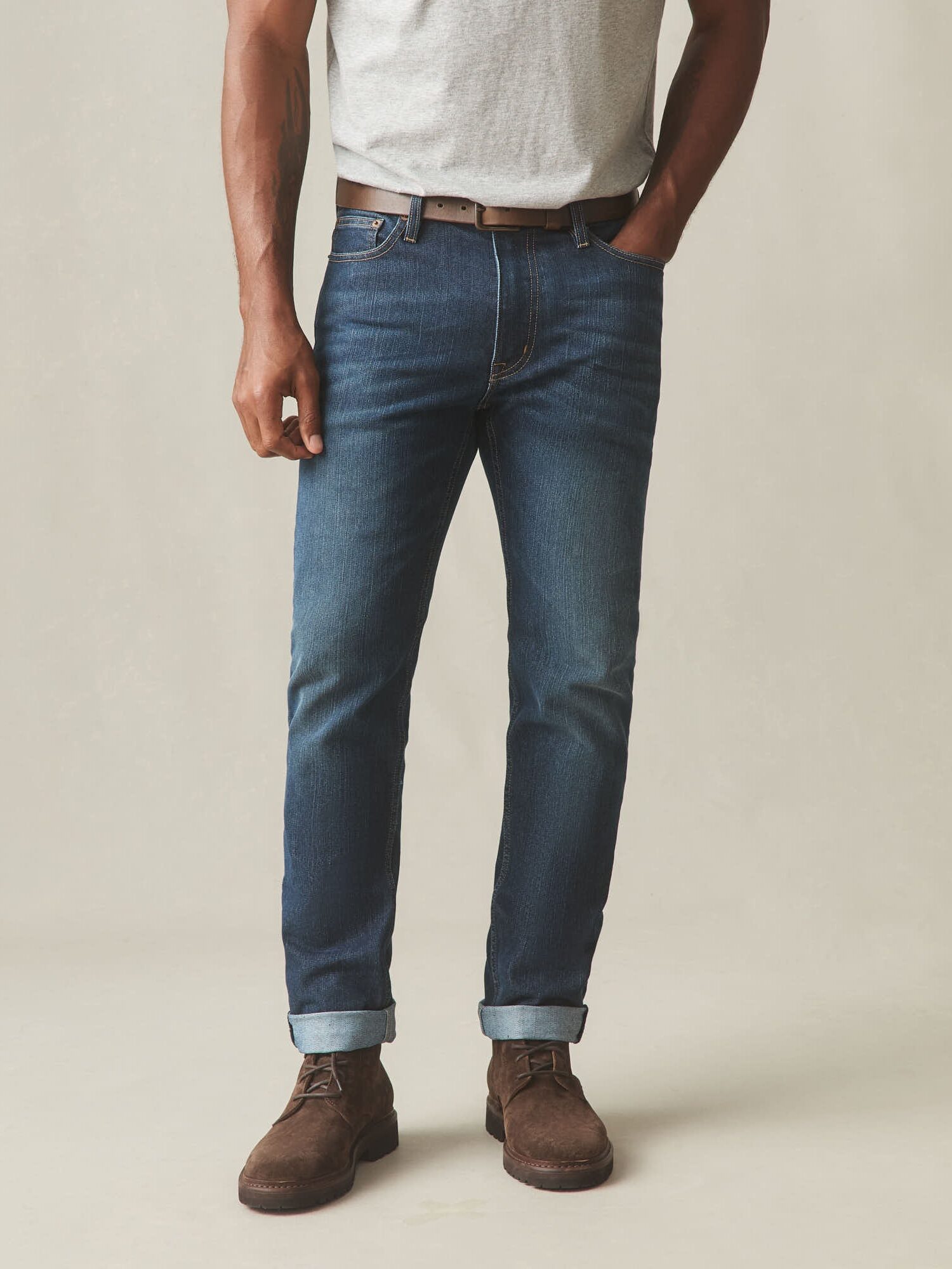 A person wearing a light gray t-shirt, blue cuffed jeans, a brown belt, and brown suede boots stands with one hand in their pocket against a neutral background.