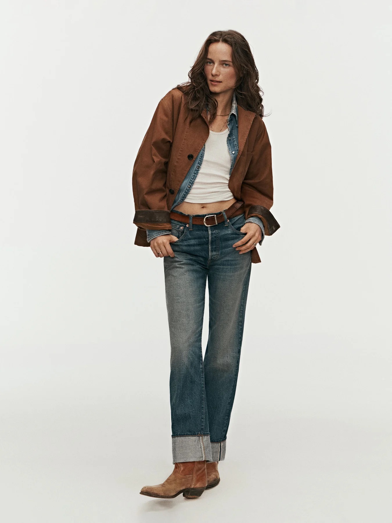 A person stands against a plain background wearing a brown jacket, white tank top, blue jeans with cuffs, a belt, and brown boots.