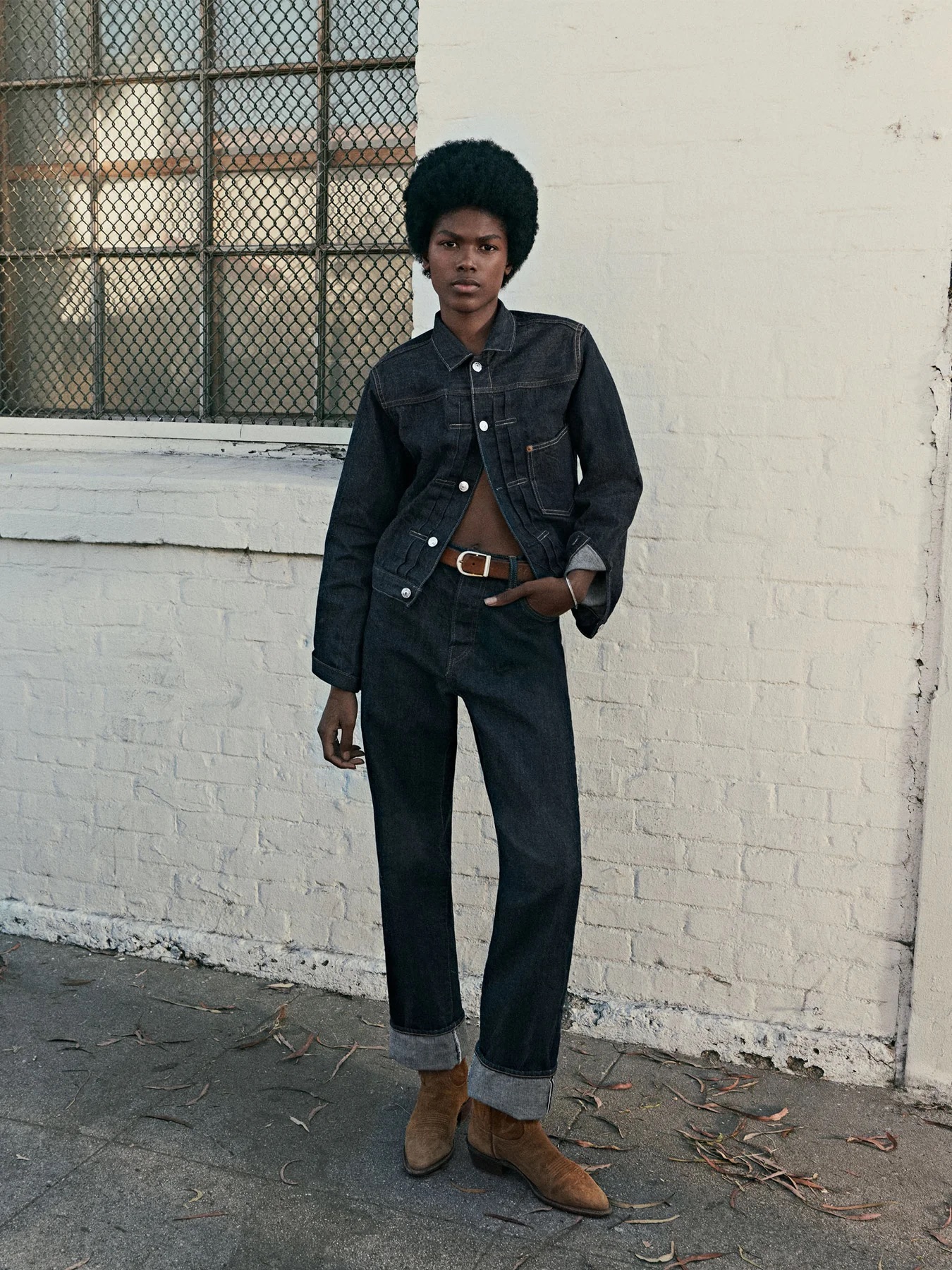 A person stands outdoors against a white brick wall, wearing a dark denim jacket and jeans, a brown belt, and brown boots.