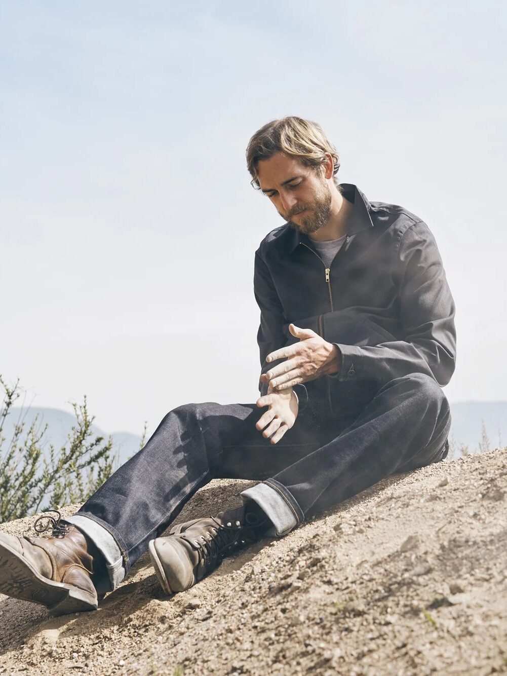 A man sits on a sandy slope outdoors, brushing sand off his hands, wearing a dark jacket, jeans, and brown boots. Shrubs and a hazy sky are in the background.