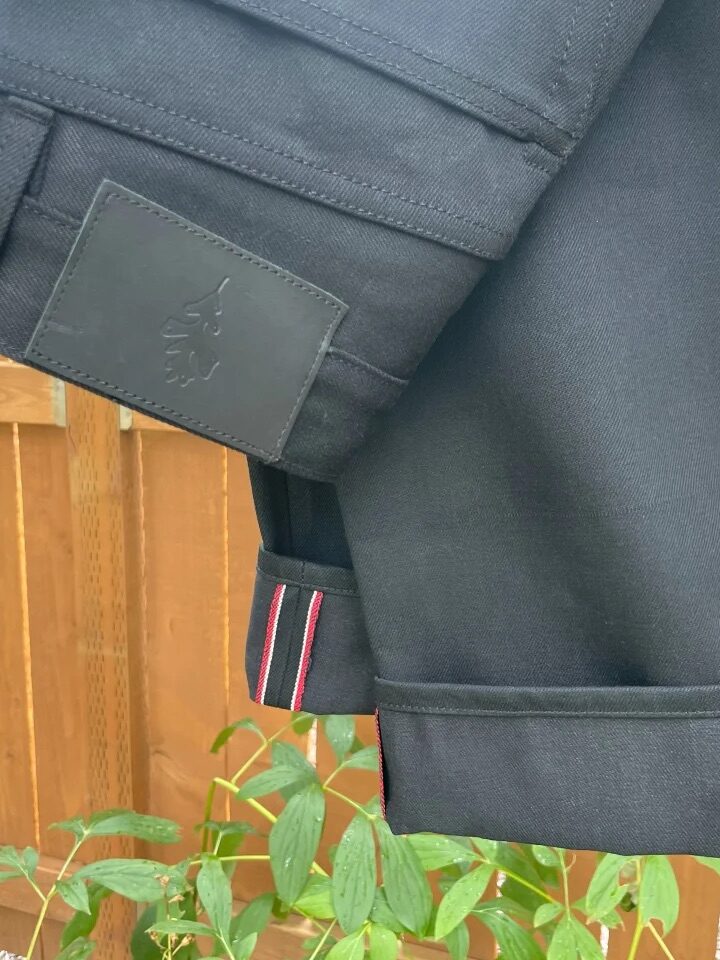 A pair of black pants with a black patch and red and white stitching near the hem is draped over a wooden fence above green plants.