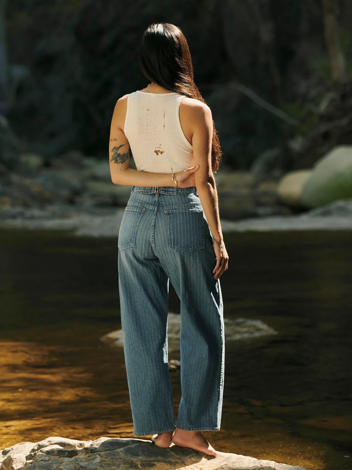 A person with long dark hair, wearing a sleeveless white top and blue jeans, stands barefoot on rocks beside a river, facing away from the camera.