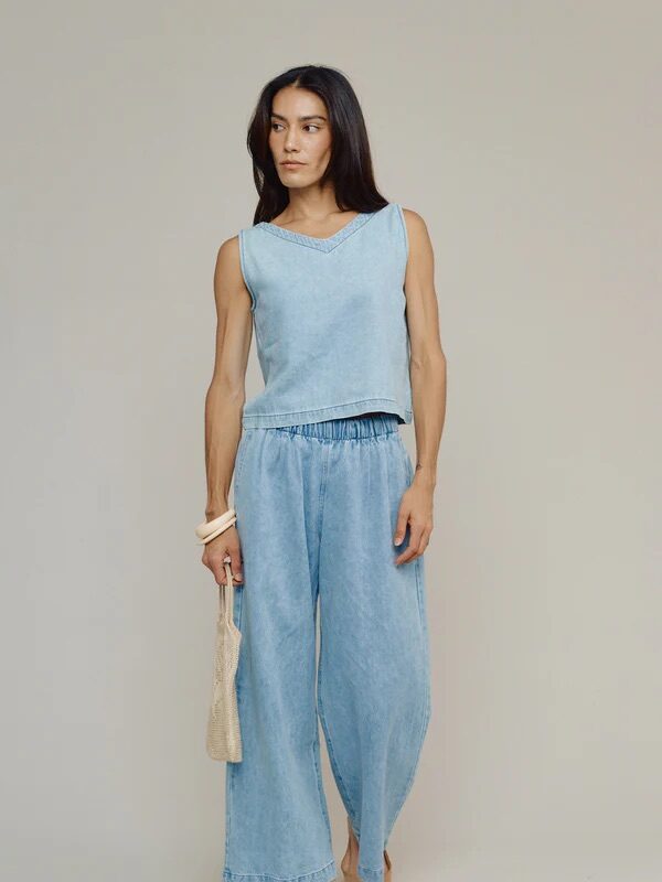 A person wearing a sleeveless light blue denim top and matching wide-leg pants holds a small beige bag, standing against a plain background.