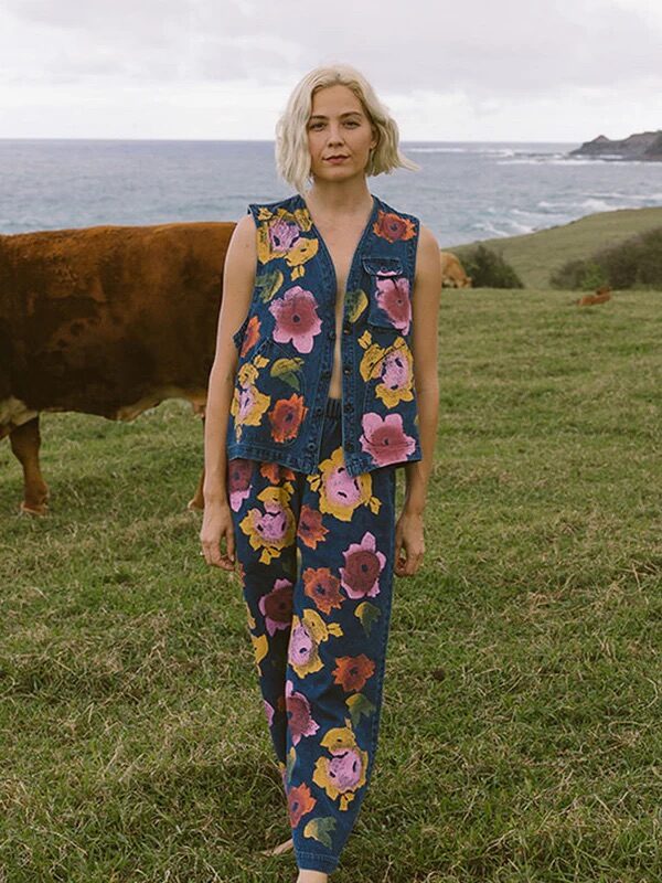 A woman with blonde hair stands barefoot on grass near the ocean, wearing a blue sleeveless denim outfit with a large pink and orange floral print. A brown cow is in the background.