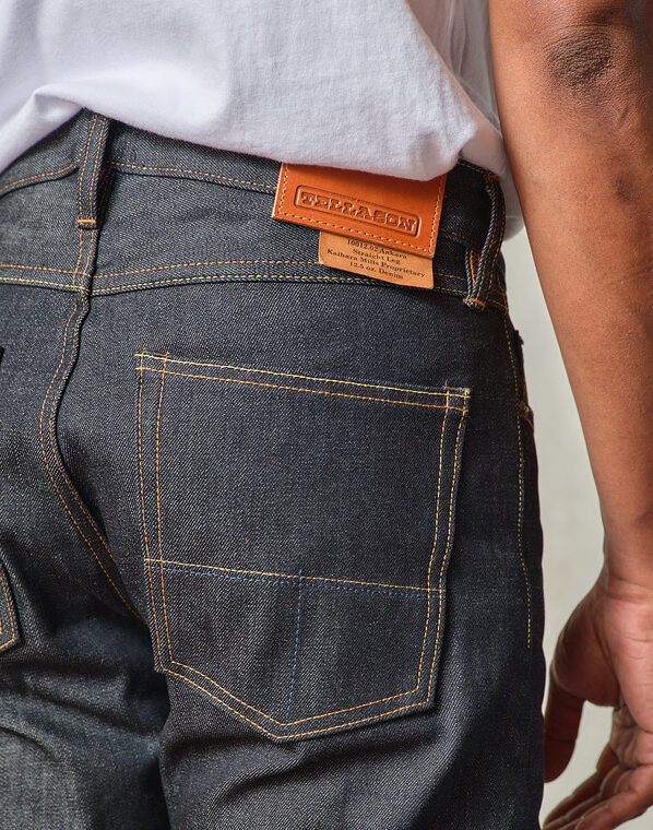 A person wearing dark denim jeans and a white shirt, shown from the back waist down, with a leather brand label on the jeans.