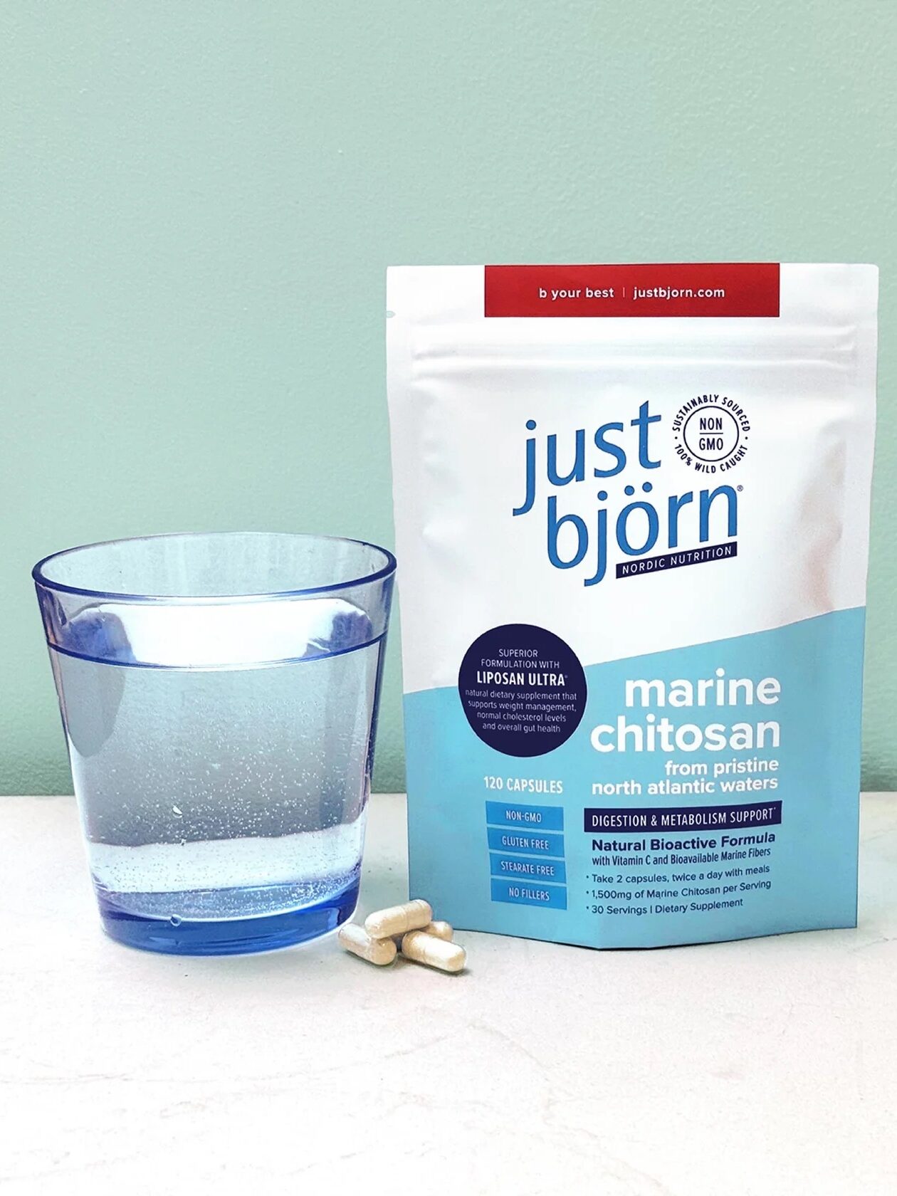 A glass of water, three capsules, and a packet labeled "just björn marine chitosan" dietary supplement are placed on a light surface against a pale green background.