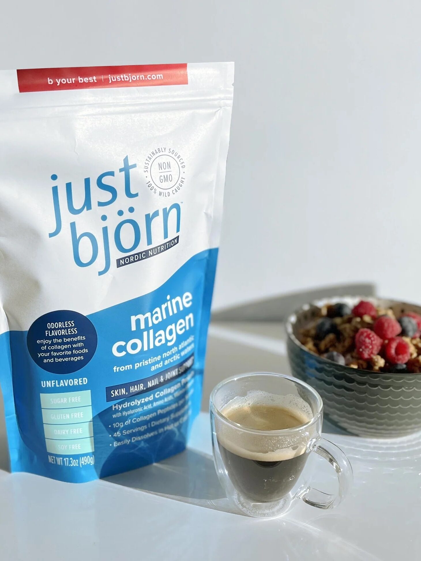 A bag of just björn marine collagen stands next to a glass cup of espresso and a bowl of granola topped with raspberries and blueberries on a white surface.