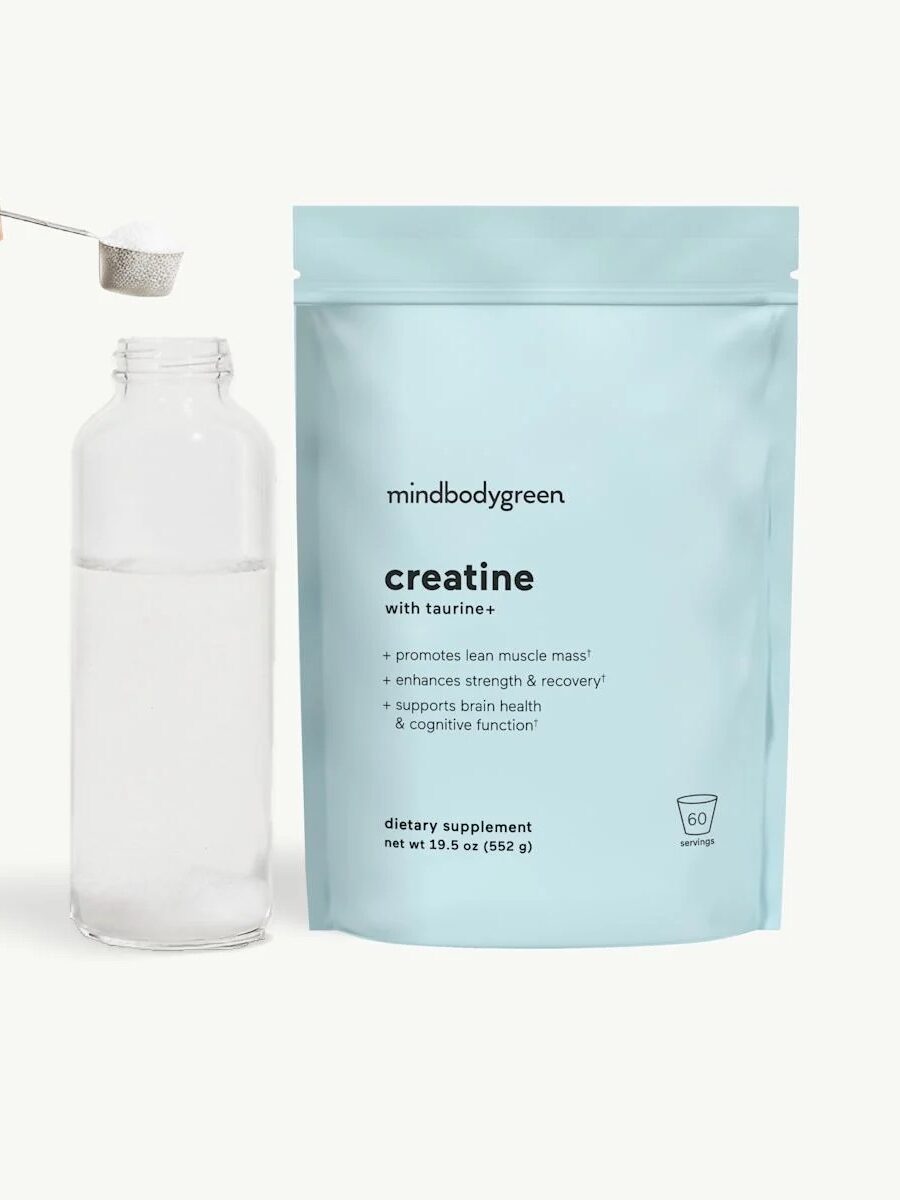 A hand adds a scoop of creatine powder to a glass bottle of water next to a blue pouch labeled "mindbodygreen creatine.