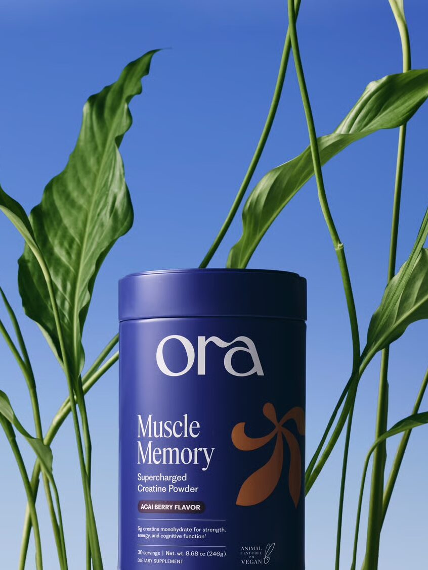 A container of Ora Muscle Memory creatine powder stands in front of green plant leaves against a blue sky background.