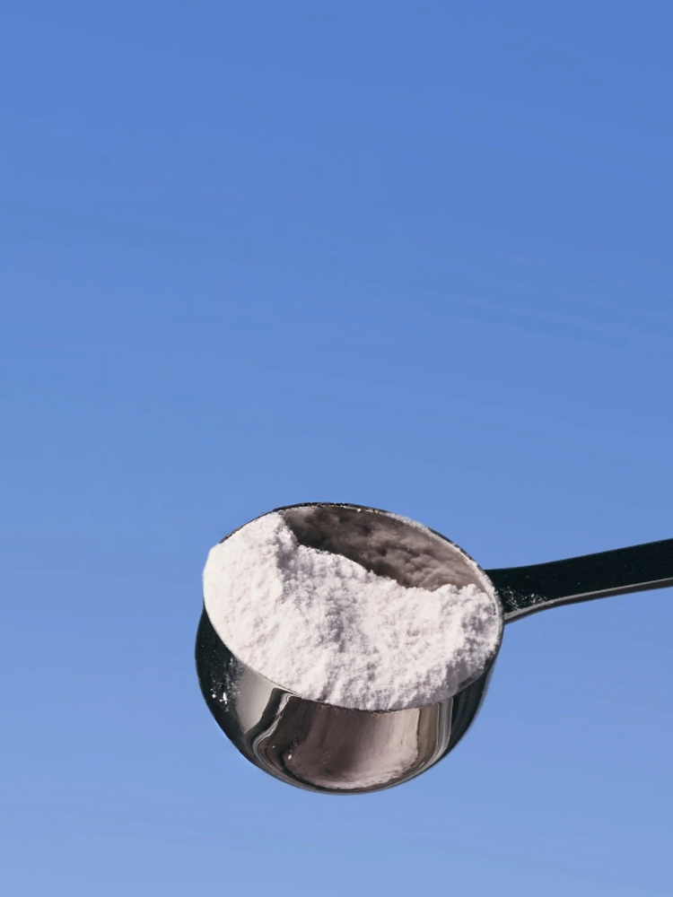A metal scoop filled with white powder is held up against a clear blue background.
