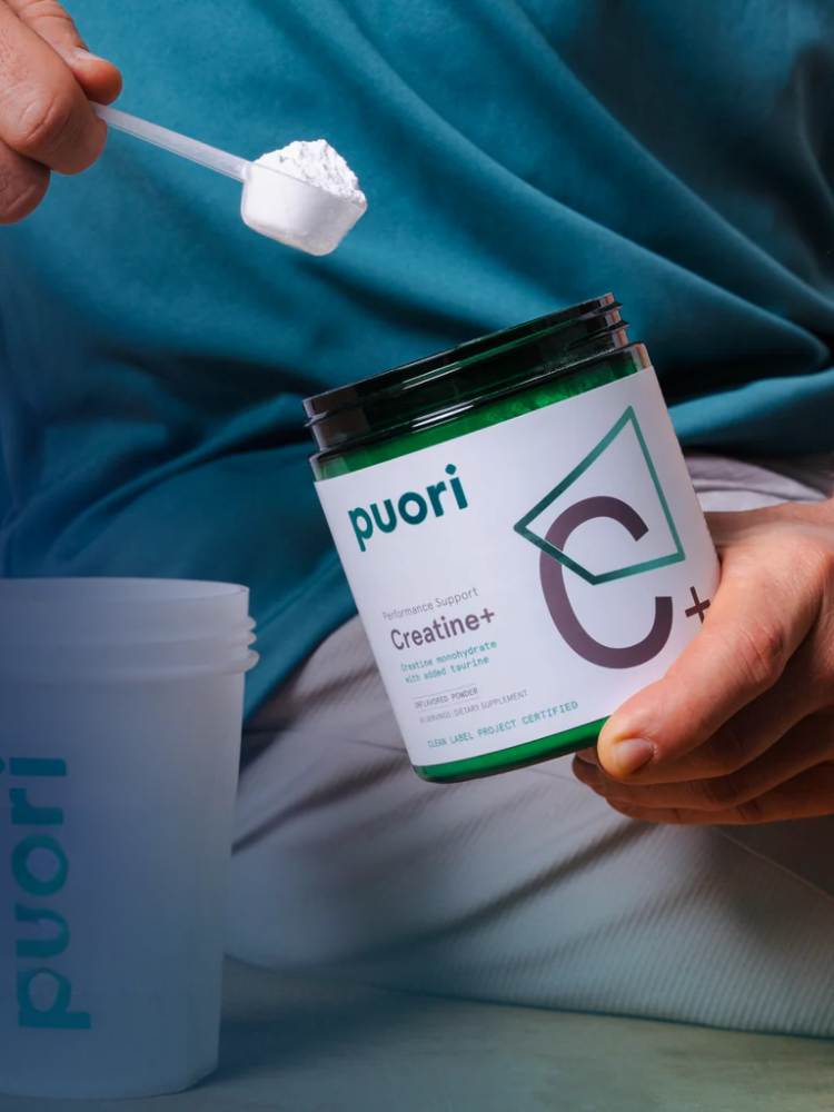 A person scoops creatine powder from a jar labeled "Puori Creatine+" next to a translucent shaker bottle with the same branding.