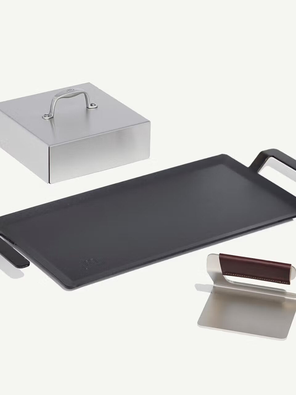 Rectangular black griddle with handles, stainless steel press with handle, metal spatula with brown handle, and a metal scraper on a white background.