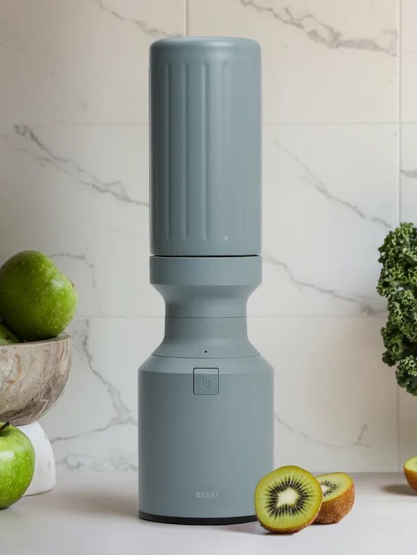 A gray countertop blender stands upright, surrounded by green apples, kiwis (whole and sliced), and a bunch of kale on a white surface with a tiled background.