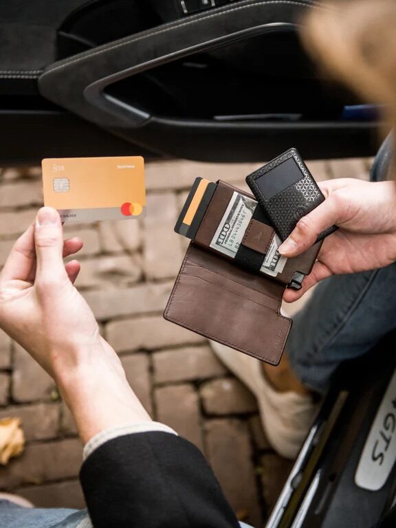 A person seated in a car holds an open wallet and a yellow Mastercard debit card, with credit cards and cash visible.