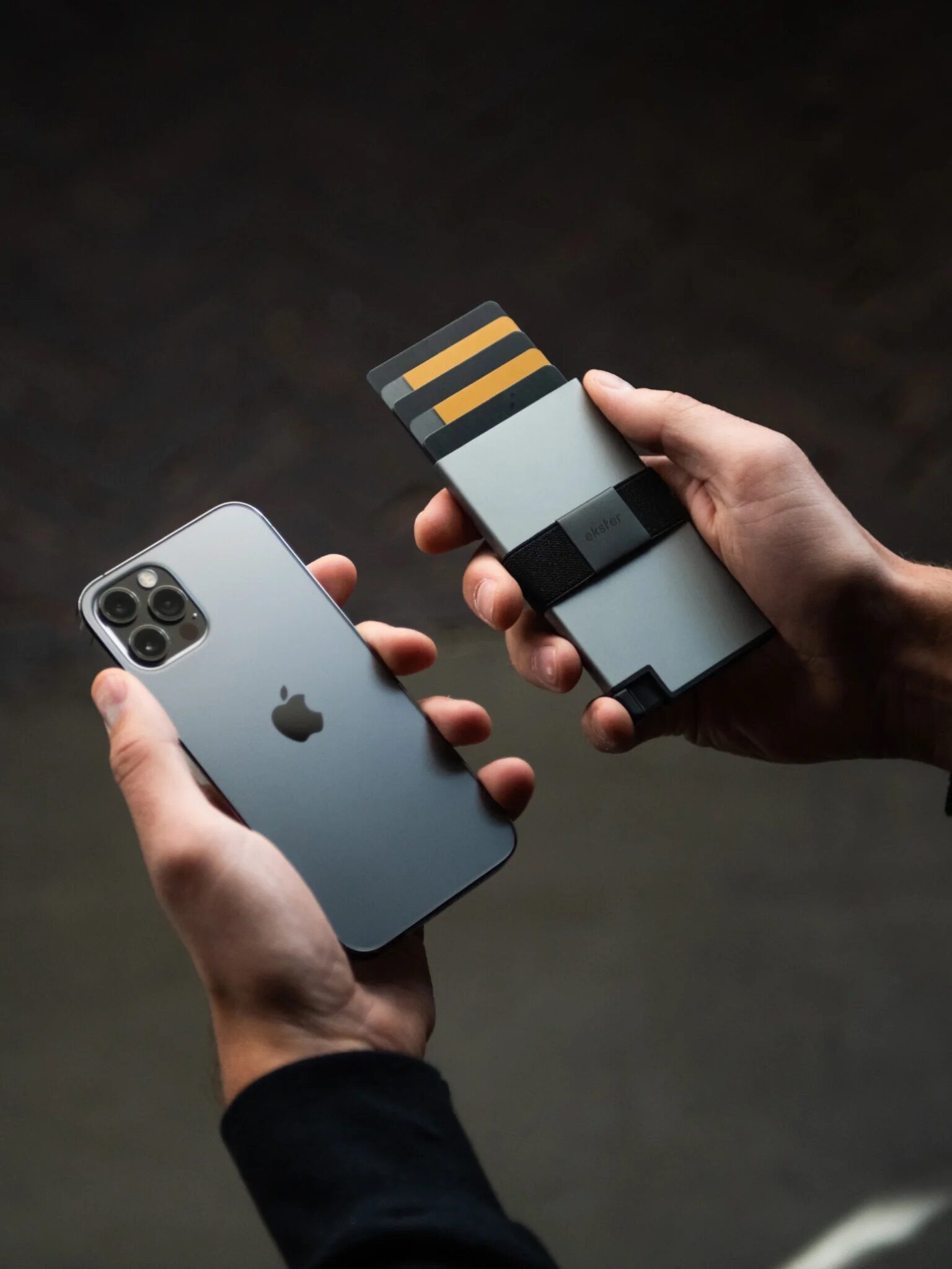 A person holding a silver iPhone in one hand and a modern, minimalist cardholder with several cards in the other hand.