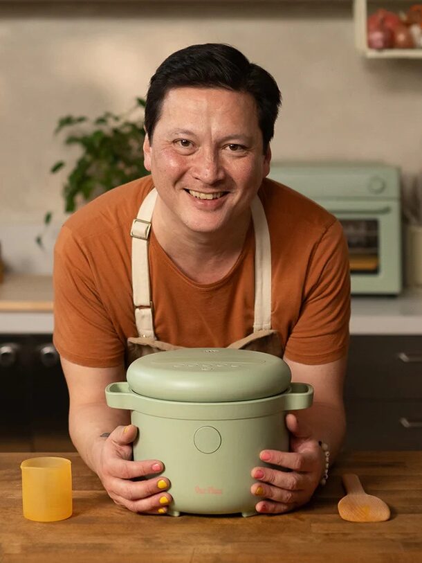 A person in an orange shirt and apron smiles while holding a green cooking pot on a kitchen counter with utensils and ingredients in the background.