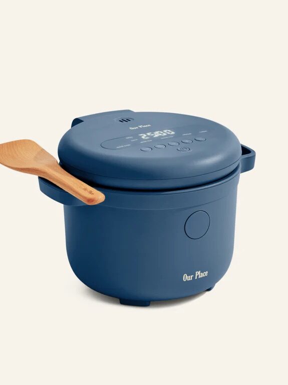 A blue electric rice cooker with a digital display, lid, side handles, and a wooden spatula resting on top, labeled "Our Place.