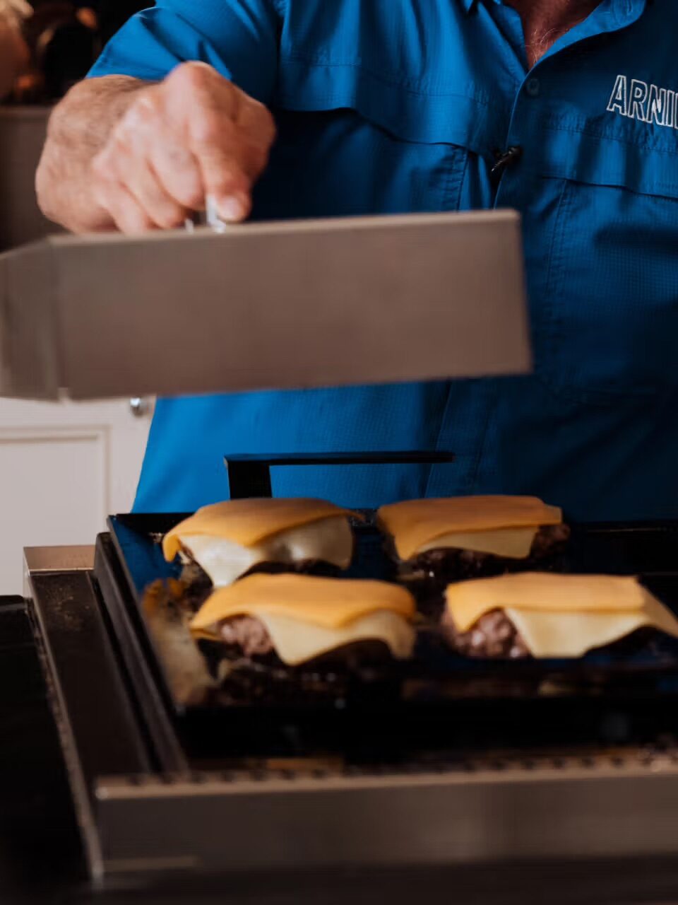 A person in a blue shirt cooks cheeseburgers on a griddle, using a large metal spatula to press down on the burgers.