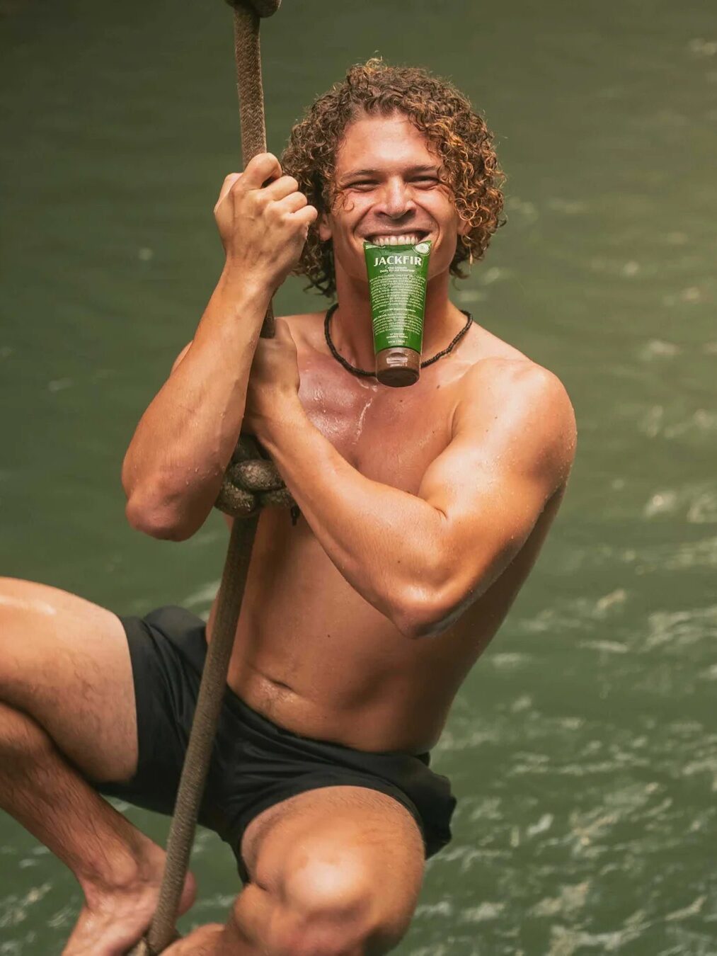 A smiling shirtless man in black swim trunks hangs from a rope over water, holding a green bottle in his mouth.