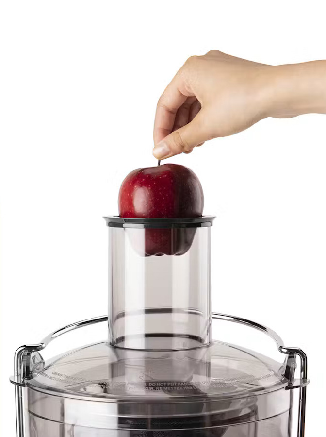 A hand places a red apple into the feed chute of a juicer against a white background.
