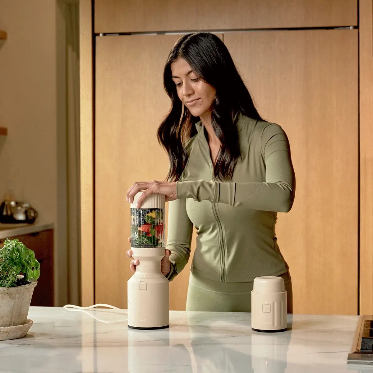 A woman in a light green outfit uses a portable blender with fruits and vegetables inside on a kitchen counter.