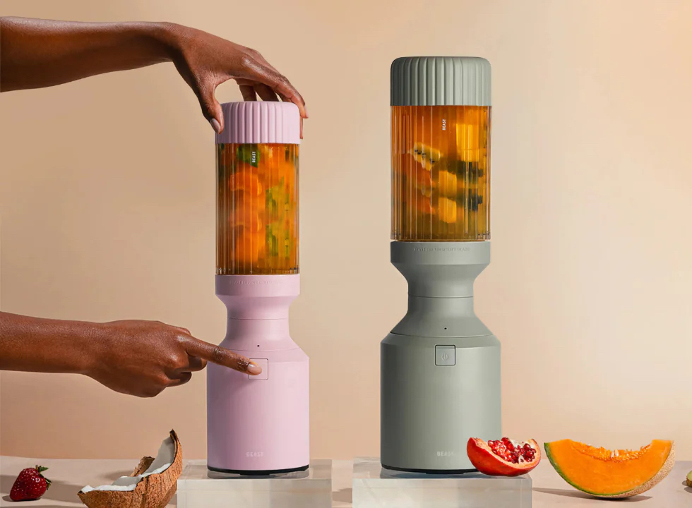 Two hands operate pink and gray portable blenders filled with chopped fruit, set on a surface with fruit pieces around them against a beige background.