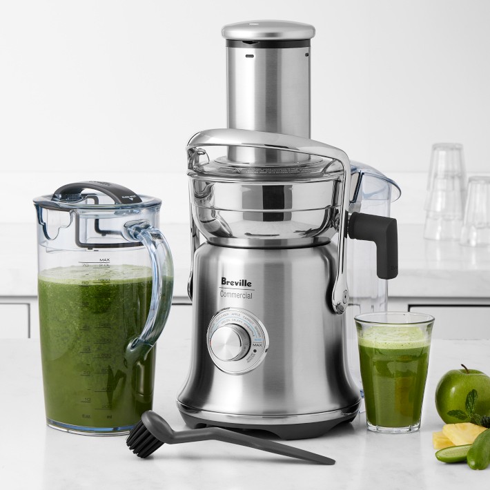 A stainless steel Breville juicer on a countertop with a full pitcher and a glass of green juice, a cleaning brush, an apple, and lemon slices nearby.