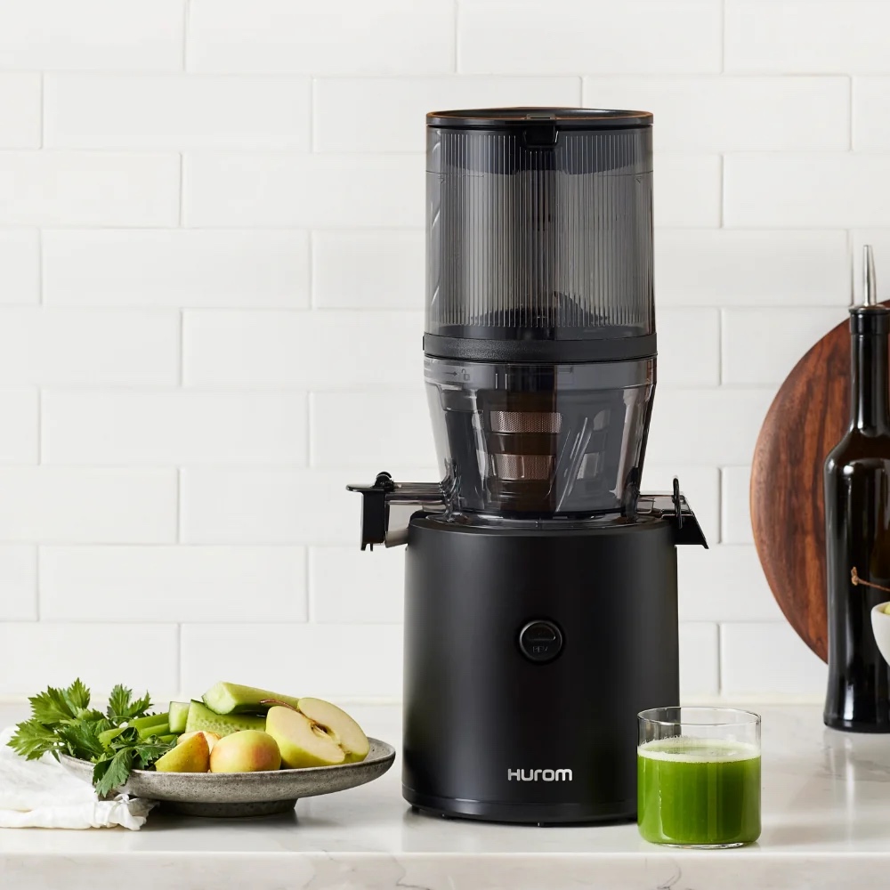 A black Hurom juicer on a kitchen counter with a plate of apples and celery, and a glass of green juice beside it.