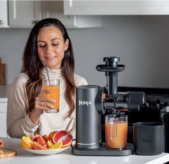 A woman sits at a kitchen counter, holding a glass of orange juice next to a Ninja juicer and a plate of sliced citrus fruits.