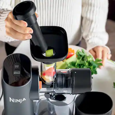 Person using a Ninja food processor to push a piece of produce into the chute, with other vegetables and greens on the counter nearby.