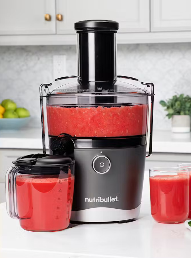 A Nutribullet juicer filled with red juice, a matching pitcher, and two glasses of juice on a white kitchen counter.