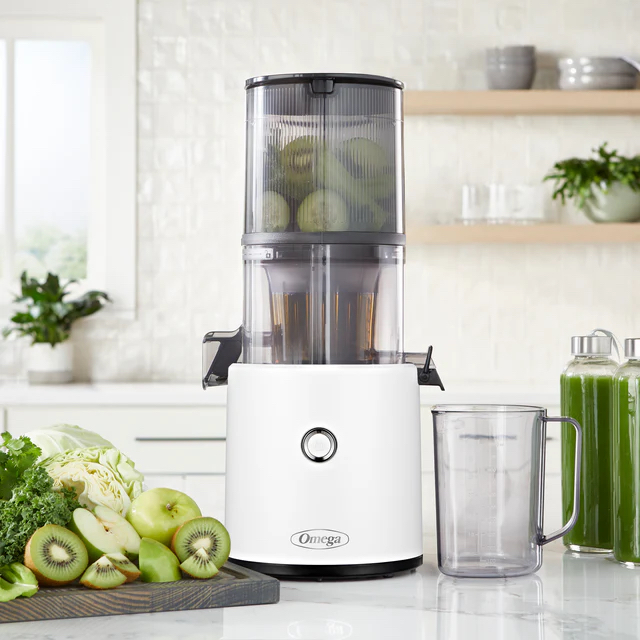 A white Omega juicer with a container of whole kiwis is on a kitchen counter, surrounded by chopped fruits, greens, and two jars of green juice.