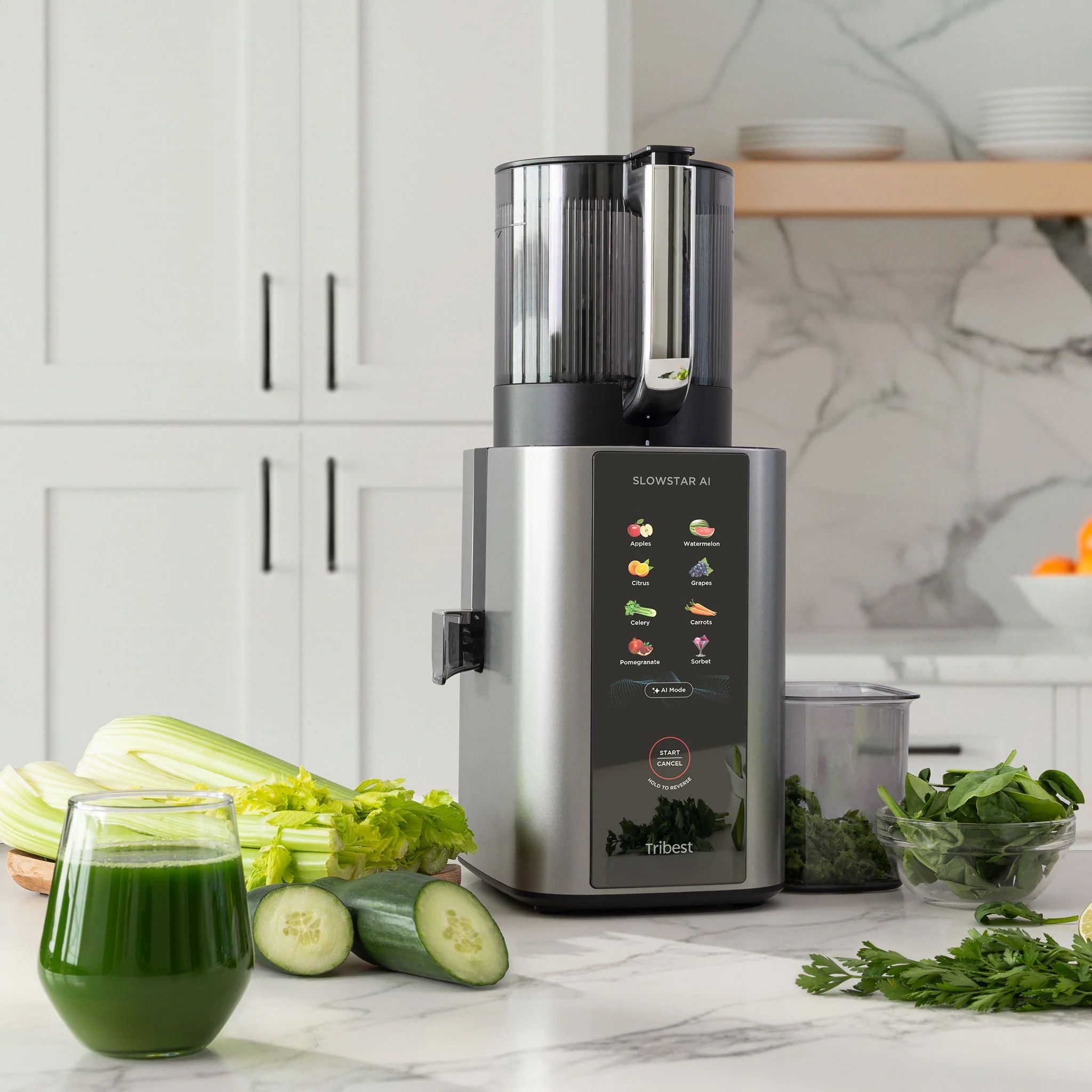 A slow juicer sits on a kitchen counter surrounded by celery, cucumber, greens, and a glass of green juice, with kitchen shelves and a plant in the background.