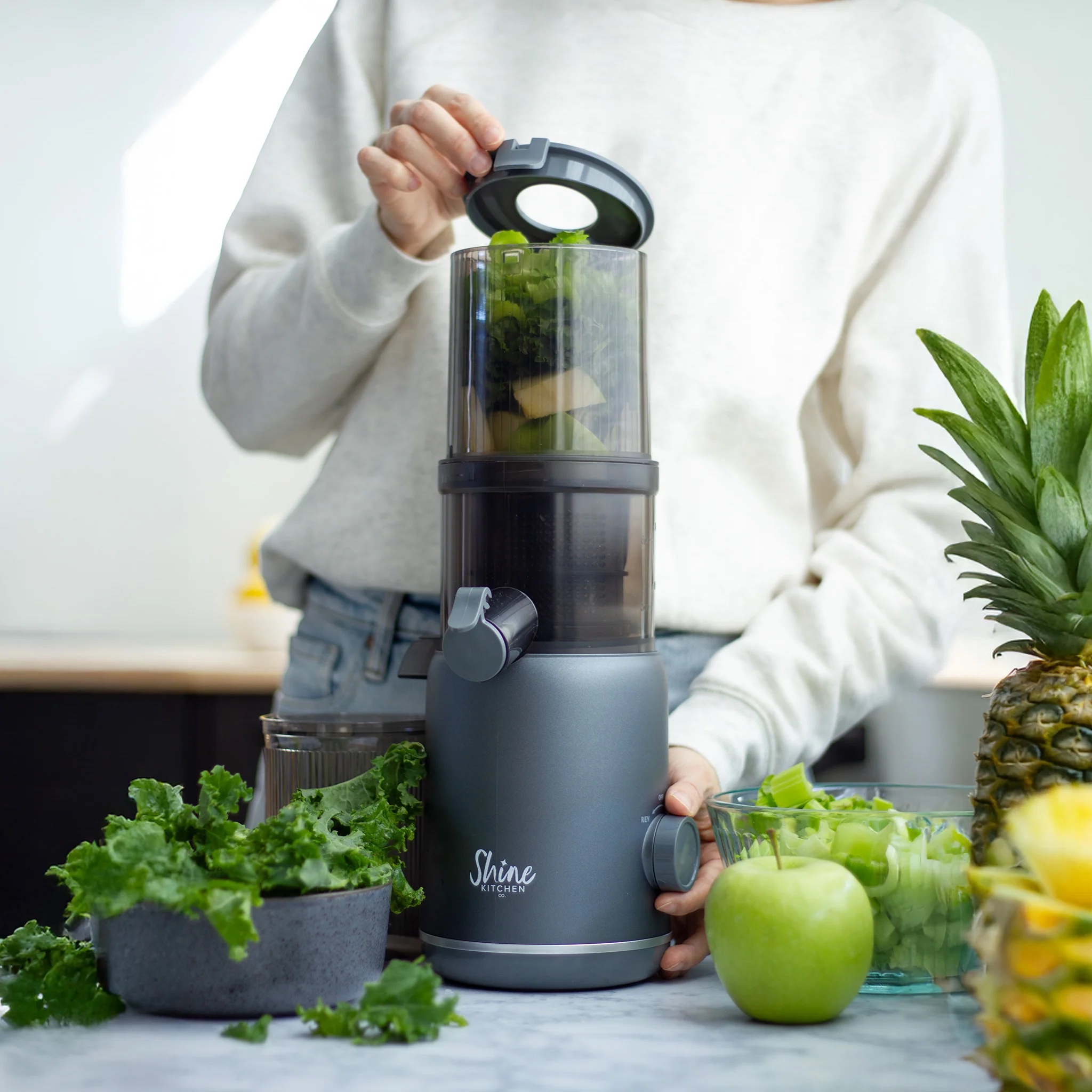 Person using a Shine Kitchen Co. blender filled with green vegetables, surrounded by kale, celery, an apple, and a pineapple on a kitchen counter.