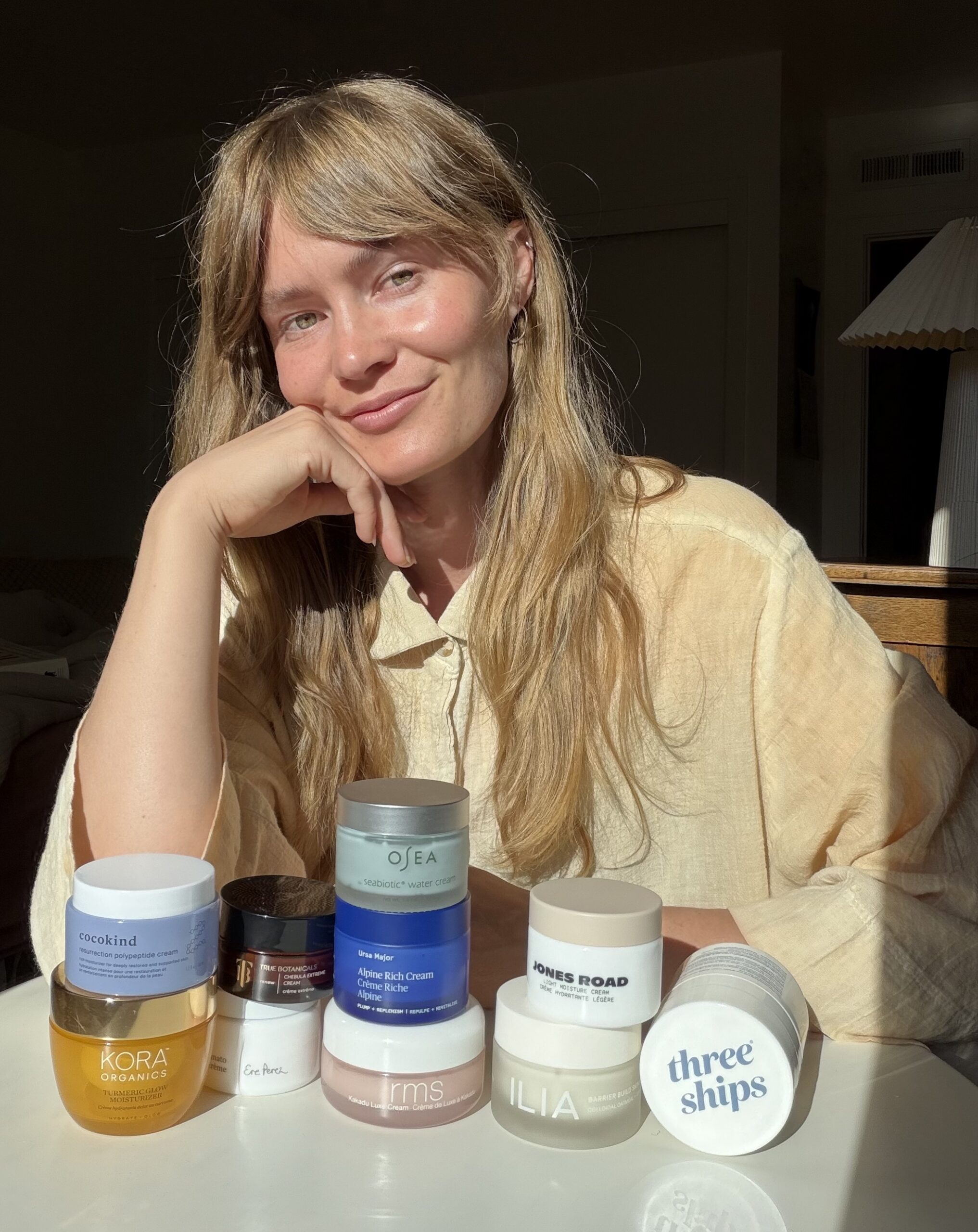 A person with long blonde hair sits at a table, smiling, resting their chin on their hand, with several skincare jars and containers arranged in front of them.