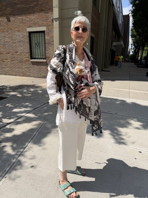 Older woman with short gray hair, sunglasses, and teal sandals stands on a sunny sidewalk, wearing white pants, a patterned shawl, and holding a small dog wrapped in a blanket.