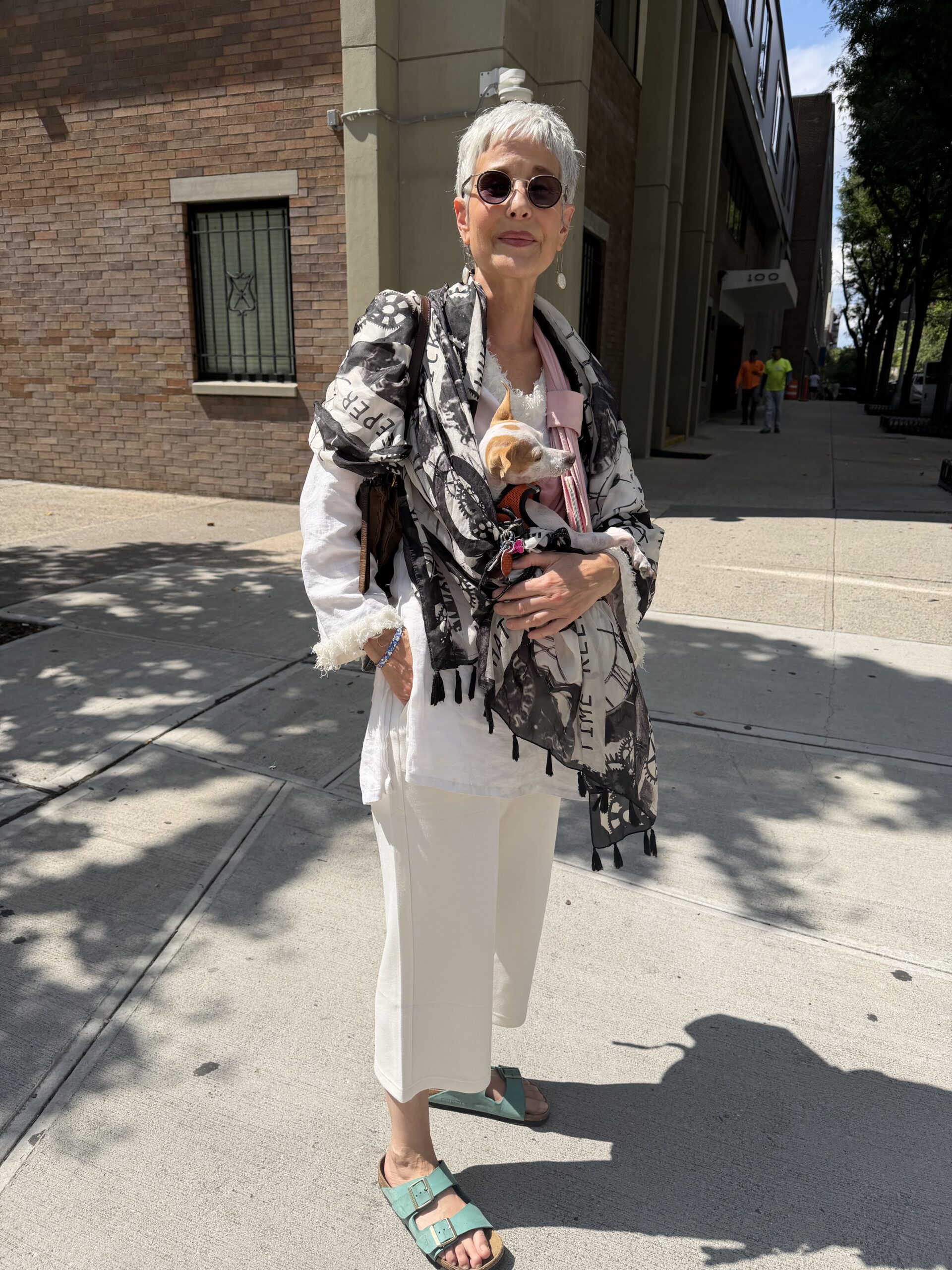 Older woman with short gray hair, sunglasses, and teal sandals stands on a sunny sidewalk, wearing white pants, a patterned shawl, and holding a small dog wrapped in a blanket.