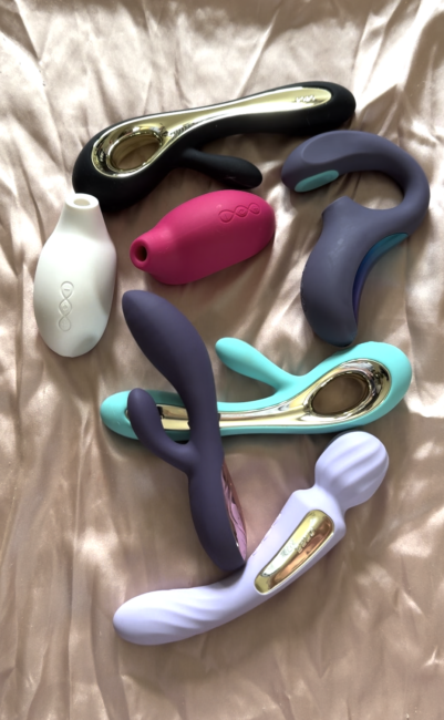Six different silicone vibrators in various colors and shapes are displayed on a light satin fabric background.