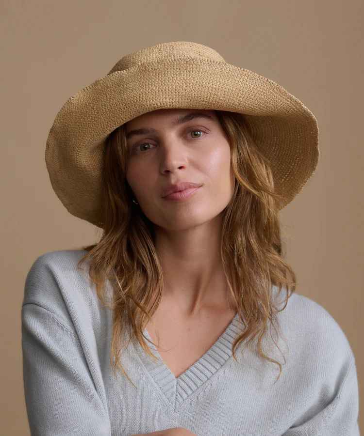 A woman with wavy light brown hair wears a wide-brimmed straw hat and a light gray v-neck sweater, posing against a plain beige background.