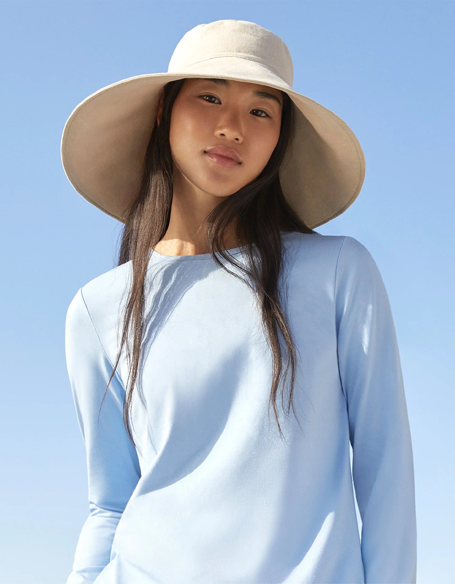 Woman wearing a wide-brimmed beige hat and light blue long-sleeve shirt stands outdoors against a clear blue sky.