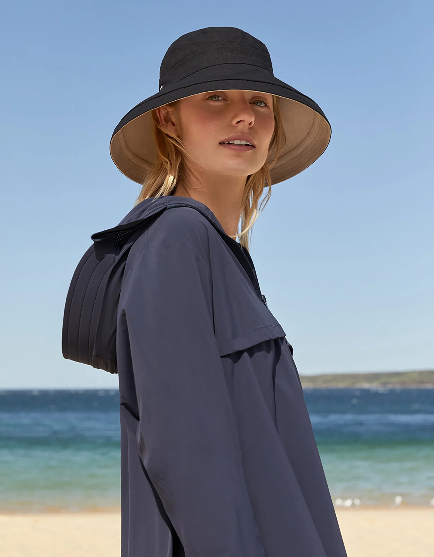 Woman wearing a wide-brimmed black hat and navy jacket stands on a sandy beach with blue water and a clear sky in the background.