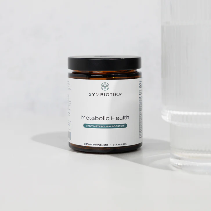 A jar of Cymbiotika Metabolic Health dietary supplement capsules is placed next to a clear glass of water on a light surface.