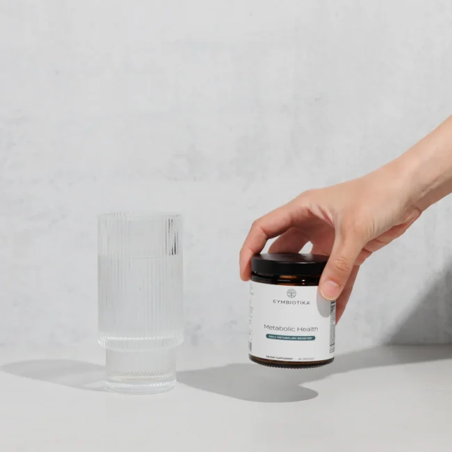 A hand holds a jar labeled "Metabolic Health" next to a glass of water on a light grey surface.