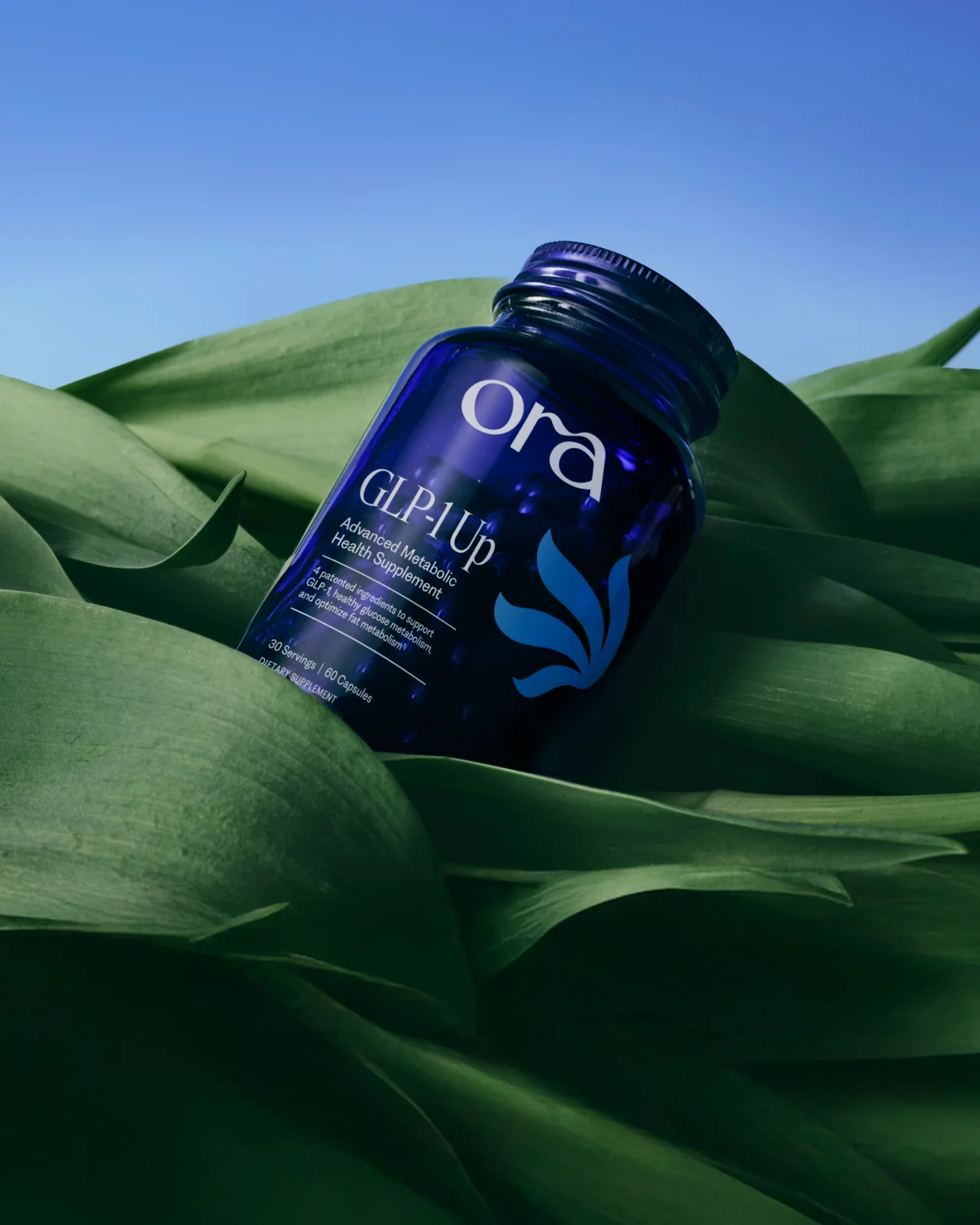 A blue Ora GLP1 Up supplement bottle rests on large green leaves against a blue sky background.