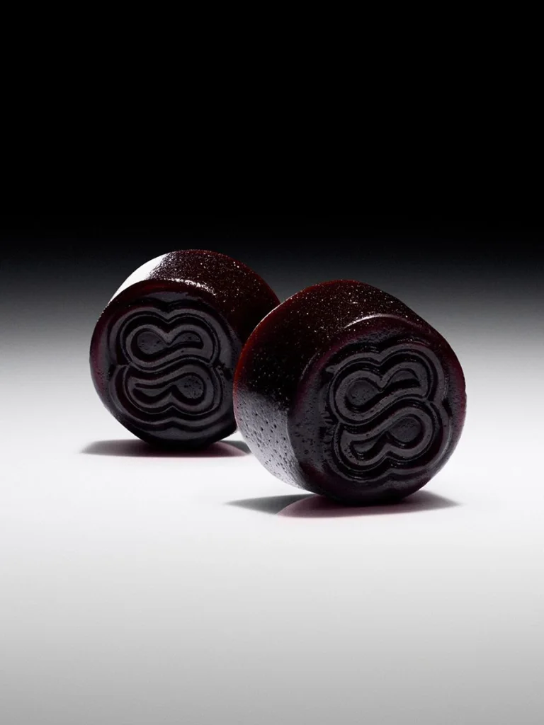 Two round, dark-colored candies with a shiny surface and embossed swirl patterns are positioned on a white surface against a dark gradient background.