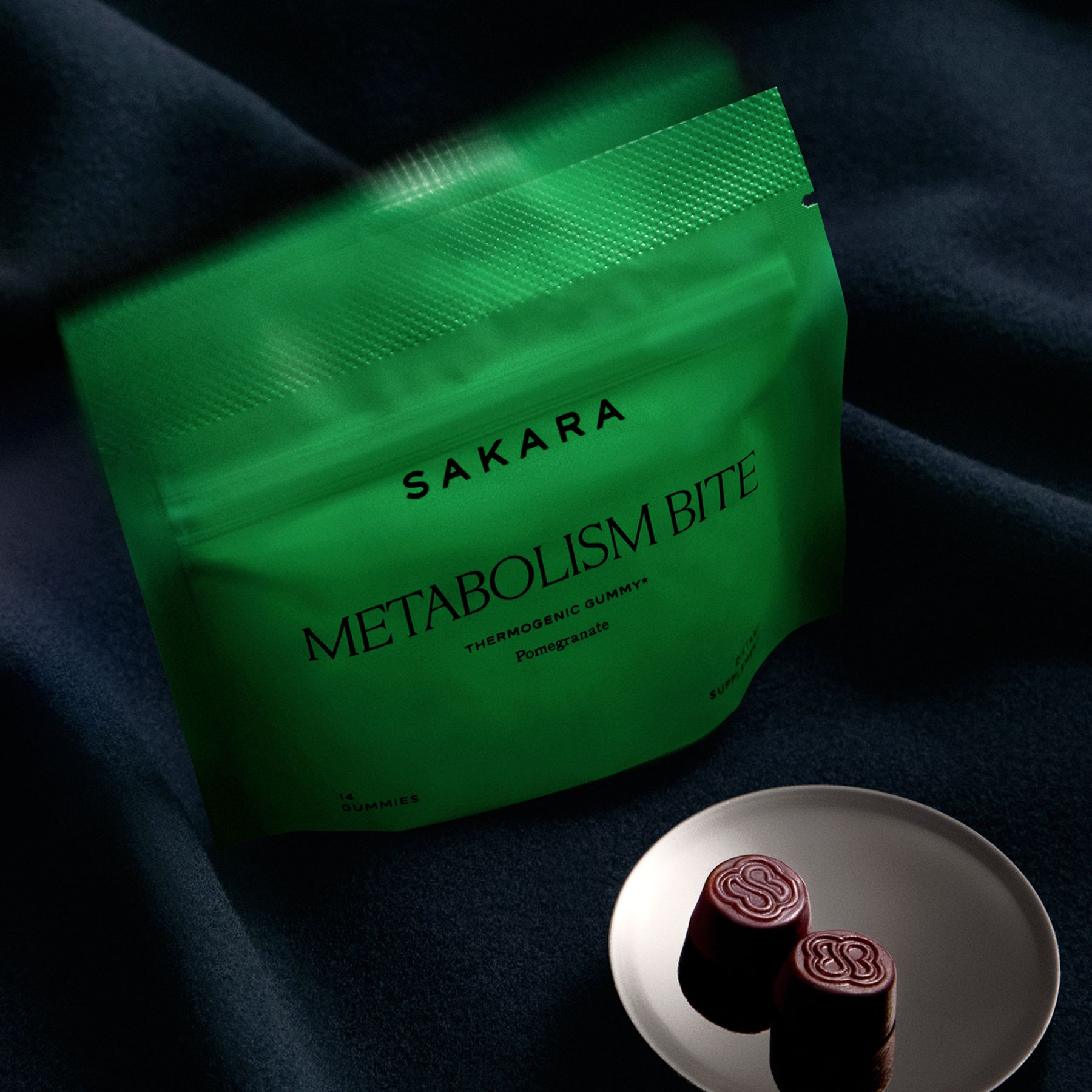 A green package labeled "Sakara Metabolism Bite" sits on a dark cloth next to a small plate holding two round, dark gummies embossed with an "S".